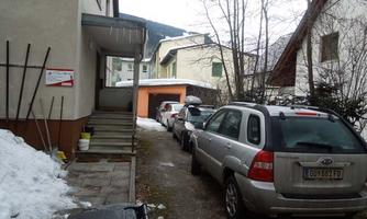 A narrow driveway with parked cars next to a residential building. In the background, snow-covered areas and trees are visible.