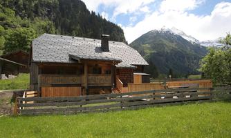 A traditional wooden house in a green landscape. In the background, mountains and a clear sky are visible.