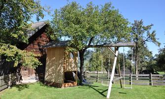 A beautiful yard with a wooden house and a swing set. In the background, a large tree and a meadow can be seen.