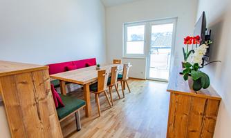 A bright and modern dining area with a wooden table and chairs. A large window front provides plenty of light and access to a balcony.