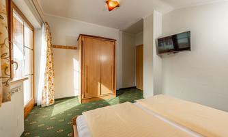 A simple bedroom with a double bed, a wardrobe, and a television. Large windows provide plenty of daylight.