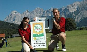 A woman and a man pose on a golf course in front of an impressive mountain backdrop. They hold a sign announcing the first Austrian organic golf course.