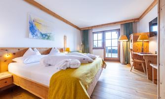 A bright and cozy bedroom with wood decor. The room features a large bed, a desk, and windows with a view outside.