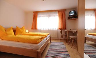 A cozy bedroom with two single beds and bright yellow bedspreads. There is a table, a chair, and a television, as well as large windows with curtains.