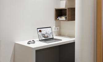 A modern workspace in a corner with a white desk and a laptop. Above, there are some shelves with books and decorative elements.