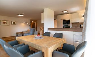 A modern dining room with a large wooden table and elegant chairs. In the background, there is an open kitchen with modern appliances.