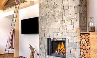 A cozy living room with a stone oven and a modern wooden shelf. Two cups of tea are on a table next to a warm blanket.