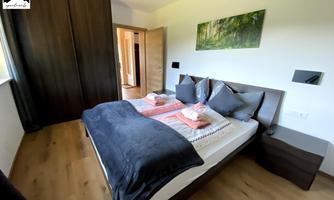 A modern bedroom with a large bed and bright wooden floors. The walls are designed minimally, and a picture of a forest hangs above the bed.