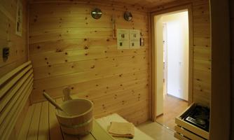 A cozy wooden sauna with benches and a bucket. In the background, a bright door is visible, opening the access to another room.