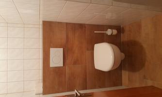 A simple bathroom with a toilet and a toilet brush holder. The walls are partially tiled, and the floor has a wood look.
