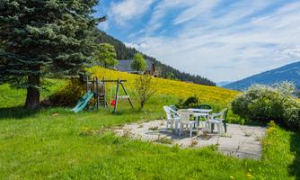 A cozy recreational area with a table and chairs on a terrace. In the background, there is a swing and a meadow with colorful flowers.