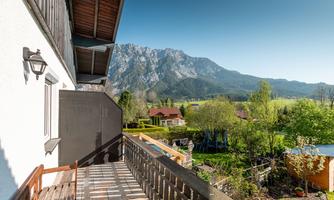 A balcony with a view of a picturesque mountain landscape. In the foreground, gardens and houses are visible.