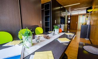 A modern dining area with a long table and colorful chairs. The decor includes plants and stylish accessories in a bright room.