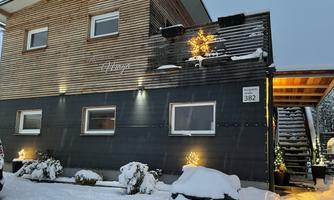A modern house with wood siding and a snow-covered ground. Small lights and Christmas decorations give the scene a festive atmosphere.