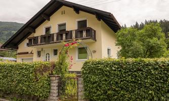 A beautiful house with a balcony and a green hedge. In the background, trees and mountains can be seen.