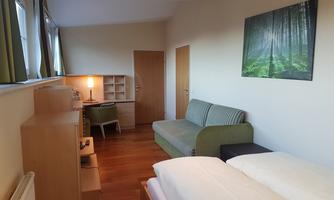 A modern bedroom with a bed, a green sofa, and a desk. The walls are painted in neutral colors and a picture of a forest hangs on the wall.
