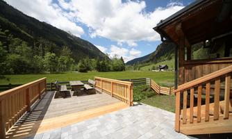 A sunny terrace with a wooden floor and a view of a green valley landscape. In the background, gentle hills and a clear blue sky can be seen.
