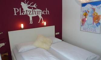 A modern bedroom with a white bed and cozy decoration. The walls are painted in a deep red and display a stylized deer as well as a piece of art titled "Austria."