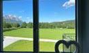 A beautiful view through a window onto a wide, green meadow and mountains in the background. The sky is clear and blue, with some white clouds.