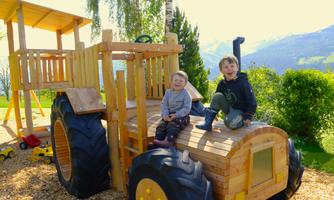 Two happy children are sitting on a wooden tractor playground. In the background, green trees and mountains can be seen.