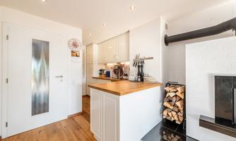 A modern kitchen with a wooden countertop and cooking area. In the background, there is a fireplace with a wood storage.