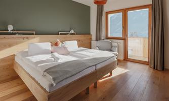 A modern bedroom with a large double bed and wood-paneled walls. The room has large windows with a view of the outdoors and cozy furniture.