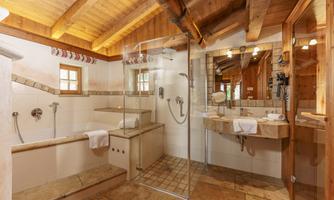 A modern bathroom with a wooden ceiling and tiled floor. It features a shower cabin, a bathtub, and a double sink.