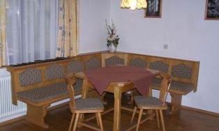 A cozy dining area with a wooden bench and a round table. The walls are decorated with pictures and there is a floral decoration at the window.