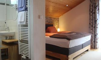 A cozy bedroom with a large bed and wood ceiling. In the background, there is a bathroom with modern amenities.