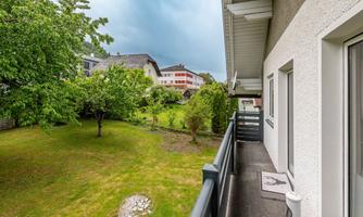 A balcony with a view of a green garden and nearby houses. The sky is cloudy and it feels calm and inviting.