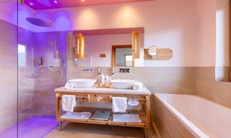 A modern bathroom with a glass shower and a bathtub. There are two sinks on a wooden shelf and soft lighting.