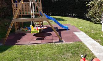 A playground in the garden with a slide, swings, and a small red vehicle. The lawn is green and the sun is shining.