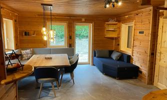 A cozy living space with wooden cladding, a large dining table, and a sofa. Large windows bring in plenty of daylight and offer access to the outdoor area.