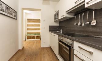 A modern kitchen with white cabinets and a dark workspace. In the background, there is a bright hallway with a visible beam and a cozy seating area.
