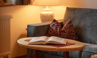 A cozy living room with a blue sofa and a round table. On the table lies an open book and a lamp provides warm light.