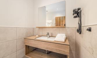 A modern bathroom with a wooden vanity and a mirror. On the vanity lies a towel and in the background, a phone is visible.