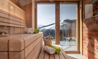 A modern sauna room with wooden benches and two towels. Through the large window, there is a view of snow-covered mountains.