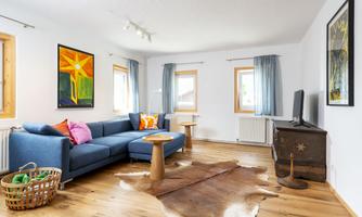 A modern living room with a blue sofa and colorful cushions. There are large windows, a wooden table, and a piece of art on the wall.