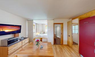 A modern room with wooden furniture and bright walls. The room contains a table, a TV wall, and a view of the bedroom.
