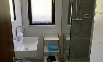A modern bathroom with a glass shower and a hand wash basin. There is also a small shelf table with towels and bath slippers.