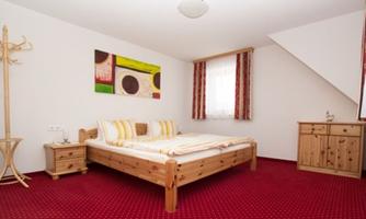 A cozy bedroom with a large bed and colorful pillows. The room has a red carpet and a small wooden piece of furniture.