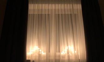 A softly illuminated window with curtains. Candlelight shines through the light fabric, creating a cozy atmosphere.