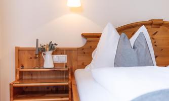 A cozy bedroom with a wooden headboard and white bedding. On the nightstand, there is a vase with flowers and a candle.