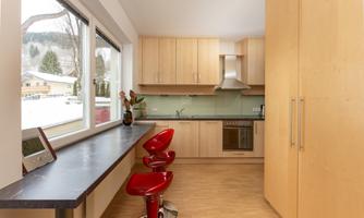 A modern kitchen with light wooden cabinets and a glass oven. Red bar stools add a stylish touch.