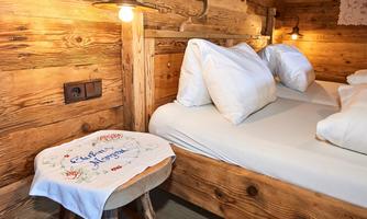 A cozy bedroom with a wooden frame bed and white pillows. Next to the bed is a small stool with a hand-embroidered cloth.