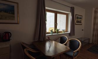 A simple dining room with a wooden table and four chairs. The window offers a view outside, and there are some plants on the windowsill.