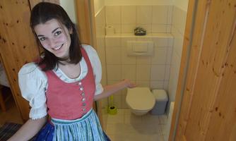 A young woman in traditional attire stands in front of a toilet. She smiles and gestures with her hand towards the room.