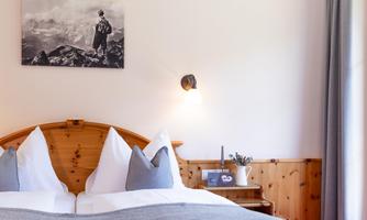 A cozy bedroom with a wooden bed and two large pillows. On the wall hangs a black-and-white picture of a mountain landscape.