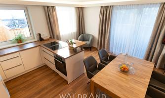 A modern kitchen with a wooden floor, bright furniture, and large windows. Comfortable chairs are around the dining table, and there is a fresh fruit bowl.