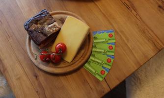 A rustic wooden board with a piece of cheese, a slice of sausage, and a small bunch of tomatoes. Next to the board are some colorful labels.